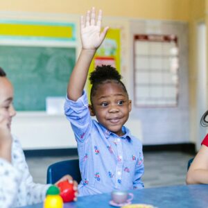 A young girl actively participates in a classroom, raising her hand to answer a question.